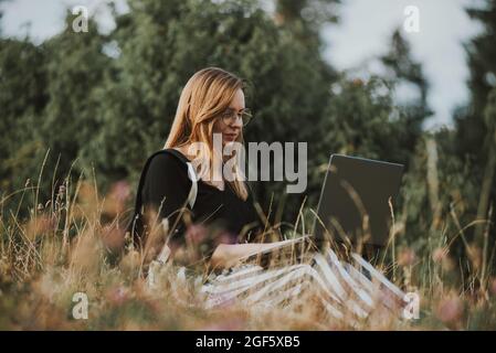 femme heureuse pigiste avec des lunettes travaillant sur ordinateur portable, à distance dans la nature Banque D'Images