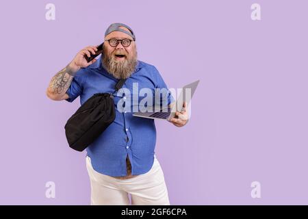 Drôle de gras homme personne avec ordinateur portable et sac de crossbody parle sur le téléphone sur fond violet Banque D'Images