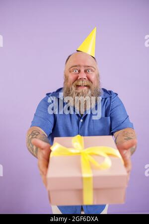 Happy FAT man dans le chapeau de fête présente boîte-cadeau debout sur fond violet Banque D'Images