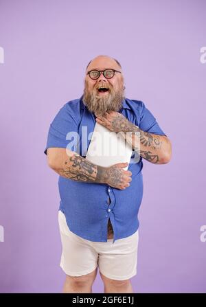 L'homme obèse à barbe excitée en chemise ajustée embrasse un ordinateur portable moderne sur fond violet Banque D'Images