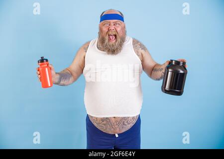 Un homme charpeux excité avec serre-tête tient des bouteilles de boisson et de supplément de protéines en studio Banque D'Images