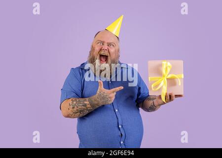 Homme excité avec un excès de poids dans des points de chemise serrés sur boîte cadeau sur fond violet Banque D'Images