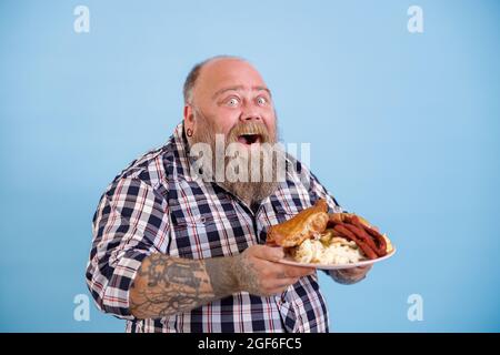 Happy Plump man tient une assiette de nourriture grasse posant sur fond bleu clair Banque D'Images