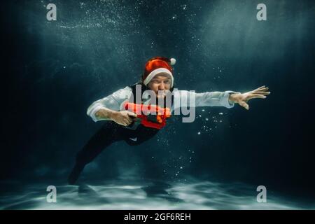Un homme sous l'eau dans un costume et un pistolet rouge dans ses mains. Le Père Noël nage sous l'eau avec un concept canon.Christmas. Banque D'Images