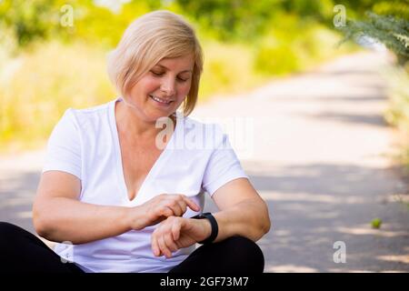 Gros plan d'une femme sportive assise avec des pieds croisés sur le chemin de la forêt, pointant sur sa montre numérique et souriant. Banque D'Images