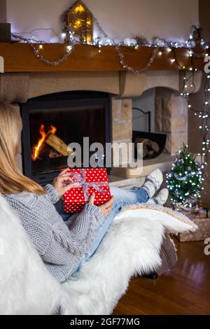Femme tenant le cadeau de noël dans les mains près de la cheminée dans l'intérieur confortable de la maison. Femme assise sur une chaise et ouvrant la boîte cadeau de noël Banque D'Images