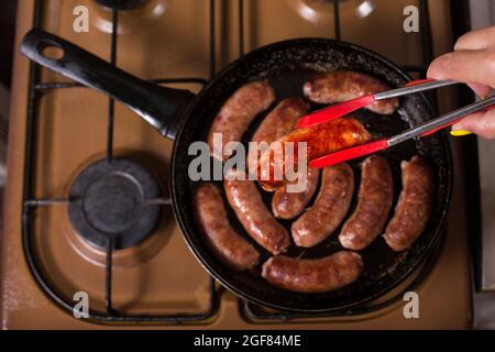 Chef cuisant des saucisses sur une poêle, vue du dessus. Hnad cueillir des saucisses dans une casserole avec des calmars de cuisine. Banque D'Images