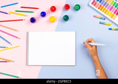 Main d'enfant avec un stylo à brossette dans un carnet de croquis d'art vierge. Vue de dessus de l'art créatif fournitures aquarelles crayons sur la table. Retour à l'école Banque D'Images