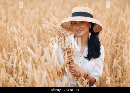 Jeune femme en chapeau de paille tenant une feuille d'épis de blé dans le champ agricole. Banque D'Images