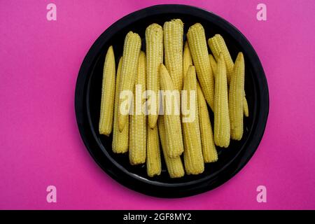 Des morceaux entiers végétaux frais et biologiques de Babycorn disposés sur une plaque noire sur fond rose plein espace vide pour écrire le texte papier peint poster Banque D'Images