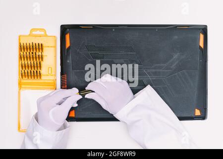 Vue du dessus des mains gantées blanches d'un ingénieur en dévissant les vis pour ouvrir le couvercle d'un ordinateur portable avant d'être réparé sur une table blanche Banque D'Images