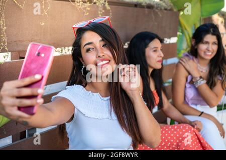 Femme latine prenant un selfie avec ses amis. Trois filles de latina ayant des boissons. Une fille hispanique avec un sourire gentil prend une photo pour ses médias sociaux. FR Banque D'Images