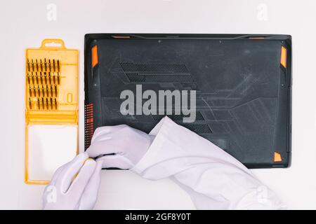 Vue du dessus des mains gantées blanches d'un ingénieur en dévissant les vis pour ouvrir le couvercle d'un ordinateur portable avant d'être réparé sur une table blanche Banque D'Images