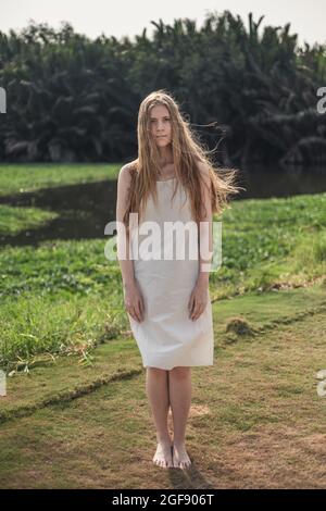 Portrait complet de la jeune jolie femme caucasienne en robe blanche debout dans le parc. Cheveux longs blonds. Beauté naturelle, sans maquillage. Banque D'Images