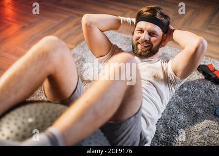 Gros plan d'un homme sportif souriant et souriant allongé sur le sol et faisant des croquantes pour les muscles abdominaux avec le fitball. Homme barbu caucasien utilisant le fitball pour l'entraînement à la maison. Banque D'Images