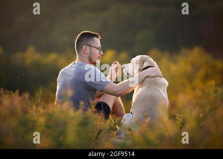 Homme avec chien assis ensemble sur la prairie. Le propriétaire d'un animal de compagnie se fait soigner pour son Labrador retriever obéissant. Banque D'Images