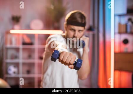 La vue avant floue de l'homme sportif actif concenté dans des vêtements de sport, debout devant l'appareil photo à la maison et faisant des coups de boxe avec des haltères. Concentrez-vous sur la main avec une haltère bleue Banque D'Images