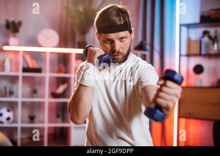 Portrait d'un homme barbu sérieux dans un serre-tête et un T-shirt décontracté qui soulève des haltères dans le salon. Le type caucasien entraîne des muscles pendant la soirée à la maison. Banque D'Images