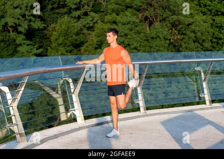 Homme sportif en vêtements de sport saisissez la cheville avec la main étirant la jambe après l'entraînement en plein air, étirez Banque D'Images