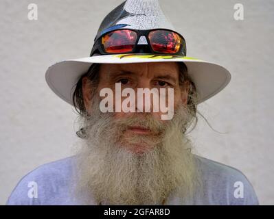 Barbu inquiet pessimiste homme caucasien âgé porte un chapeau Panama avec des lunettes de soleil colorées sur le bord et regarde le spectateur, fond blanc. Banque D'Images