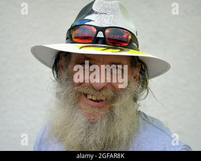 Un homme caucasien âgé optimiste et barbu sourit et porte un chapeau de Panama peint avec des lunettes de soleil colorées sur le bord de son chapeau, fond blanc. Banque D'Images