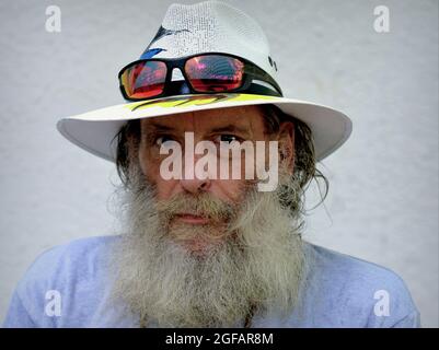 Barbu inquiet pessimiste homme caucasien âgé porte un chapeau Panama avec des lunettes de soleil colorées sur le bord et regarde le spectateur, fond blanc. Banque D'Images