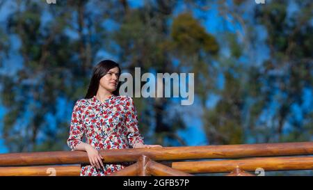 Portrait d'une belle jeune femme latine regardant à droite avec une robe à motif floral debout au milieu d'un jardin avec un fond d'arbres du Banque D'Images