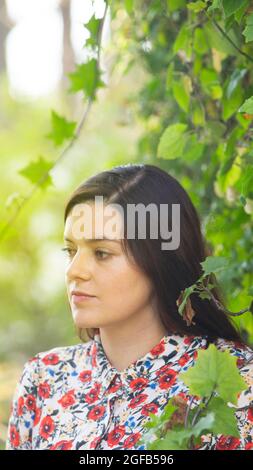 Portrait d'une belle jeune femme latine regardant à gauche portant une robe à motif floral cachée parmi une plante verte de super-réducteur pendant la matinée Banque D'Images