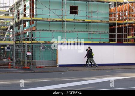 Wellington (Nouvelle-Zélande), le 25 août 2021. Un jeune couple en masque facial passe devant un chantier de construction à volets dans le centre de Wellington, en Nouvelle-Zélande, où seules les entreprises et les services essentiels peuvent fonctionner et les activités de construction ont cessé pendant que le pays est dans un strict verrouillage alors qu'il combat sa première épidémie de la variante du delta Covid-19. Crédit : Lynn grief/Alamy Live News Banque D'Images