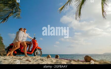 Trajet en scooter. Un couple charmant sur une moto rouge vêque de vêtements blancs sur une plage de sable. Les gens qui marchent près des palmiers tropicaux, de la mer. Location de motos. Banque D'Images