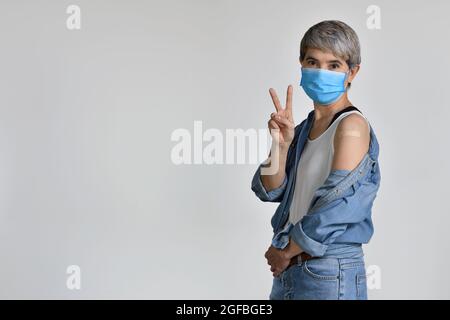 Bonne femme asiatique d'âge moyen portant un masque chirurgical pour le virus corona et vaccinée sourire avec le visage heureux à l'appareil photo faisant signe de victoire montrant un Banque D'Images