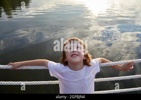 Grand angle de mignon garçon dans t-shirt blanc se pentant sur la barrière de corde et regardant vers le haut tout en se reposant près de l'eau propre de l'étang en été Banque D'Images