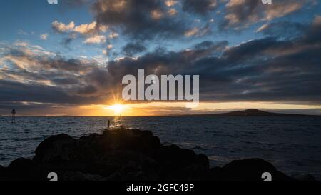 Soleil levant sur les nuages à Milford Beach. Rochers en premier plan et l'île Rangitoto au loin Banque D'Images