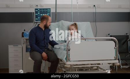 Une fille malade hospitalisée se reposant dans un lit portant un tube nasal à oxygène après avoir subi une chirurgie médicale pendant l'examen de la maladie dans la salle d'hôpital. Père expliquant le traitement de rétablissement Banque D'Images