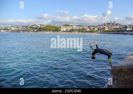 Saint-Sébastien, Espagne. 25 août 2021. Un jeune homme plonge dans l'eau de mer en effectuant un saut acrobatique à la plage de la Concha à San Sebastian.les jeunes apprécient cet été de sauter dans l'eau depuis le mur à l'extrémité de la promenade de Playa de la Concha de San Sebastian à côté de Monte Igueldo avec des sauts spectaculaires et sans le danger que les falaises de pierre offrent pour ce type de sauts. Crédit : SOPA Images Limited/Alamy Live News Banque D'Images