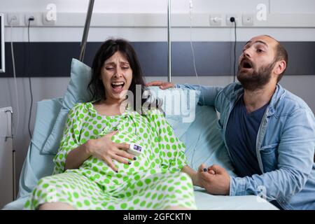 Femme enceinte entrant dans le travail ayant des contractions dans le service hospitalier à la clinique alors que l'homme nerveux hurlant pour l'infirmière et l'assistance médicale. Couple caucasien se préparant à l'accouchement Banque D'Images