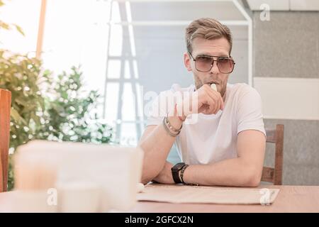 Un jeune homme avec une barbe et des lunettes de soleil fume une cigarette électronique à l'intérieur d'un restaurant ou d'un bureau Banque D'Images