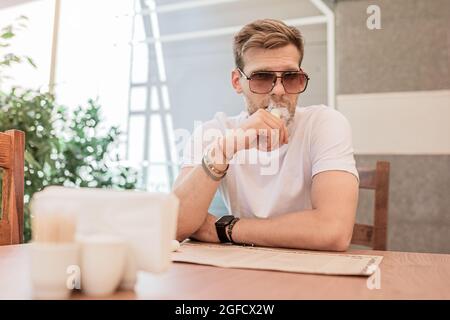 Un jeune homme avec une barbe et des lunettes de soleil fume une cigarette électronique à l'intérieur d'un restaurant ou d'un bureau Banque D'Images