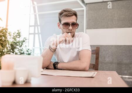 Un jeune homme avec une barbe et des lunettes de soleil fume une cigarette électronique à l'intérieur d'un restaurant ou d'un bureau Banque D'Images