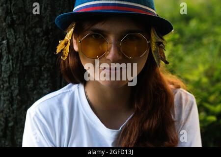 Portrait en gros plan de flou d'une jeune femme rêveuse drôle avec des cheveux bruns portant un chapeau avec des feuilles jaunes sèches et un t-shirt blanc à l'extérieur. Bonjour automne. Banque D'Images