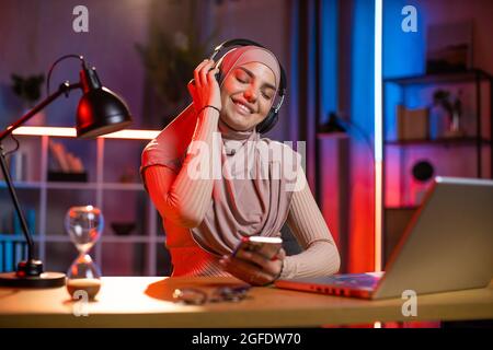 Femme charmante utilisant un smartphone et un casque pour profiter de votre chanson préférée avec les yeux fermés. Une jeune femme en hijab fait une pause tout en travaillant sur un ordinateur portable à la maison. Freelance et concept technologique. Banque D'Images