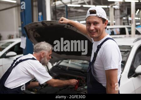 Joyeux mécanicien de voiture souriant à la caméra tout en travaillant sur une voiture avec son collègue senior Banque D'Images