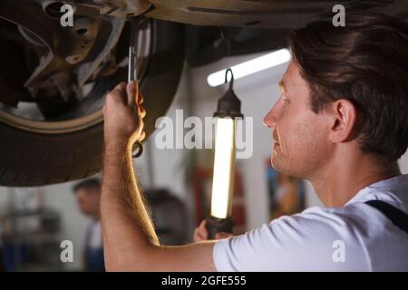 Gros plan court d'un mécanicien de voiture tenant une lampe, fixant la roue d'une auto sur un ascenseur de voiture Banque D'Images
