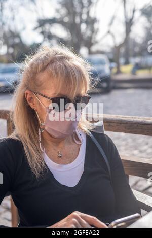 Adulte adulte mature blonde femme souriant avec des lunettes portant un masque de visage SMS au smartphone Banque D'Images