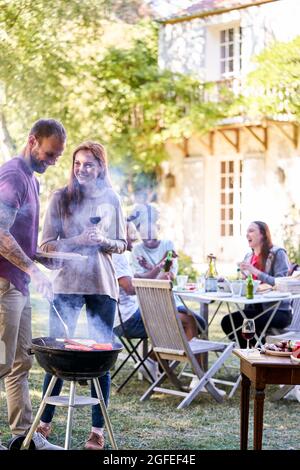 Un jeune homme prépare de la nourriture au barbecue tandis que ses amis ont des boissons en arrière-plan Banque D'Images