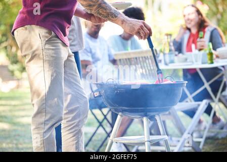 Jeune homme préparant de la nourriture au barbecue pendant que ses amis sont assis en arrière-plan Banque D'Images