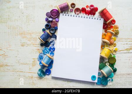 Boutons et rouleaux de spectre arc-en-ciel avec fils colorés, disposés autour d'un bloc-notes avec un espace pour le texte sur un fond en bois clair. Passe-temps et mode Banque D'Images