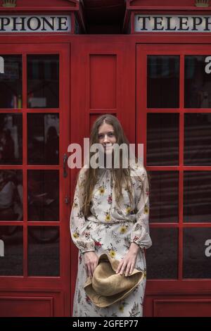 Portrait de la jeune femme caucasienne souriante en robe dans la rue à Londres. Cheveux longs et droits blonds. Cabine téléphonique rouge en anglais Banque D'Images