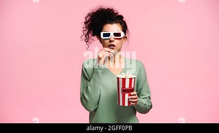 femme concentrée dans des lunettes 3d et un sweat-shirt vert mangeant du pop-corn isolé sur le rose Banque D'Images