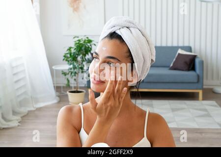 Charmante femme brune avec une serviette enveloppée sur sa tête appliquant une crème hydratante pour le visage Banque D'Images